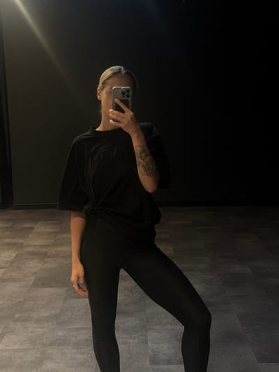 An individual is depicted indoors, capturing a mirror selfie with a light-colored smartphone that obscures their face. The subject has light-colored hair and a floral tattoo on their left forearm. They are wearing an oversized black t-shirt, tied in a knot at the front, and tight black leggings. A ring is visible on the fourth finger of their right hand. The background consists of a plain, dark-colored wall. The floor is composed of large, rectangular, gray-toned panels arranged in a staggered pattern. The lighting is subdued, with a bright reflection of an overhead light source visible in the upper left portion of the mirror. The environment presents as a minimalist indoor space, such as a fitness studio or dance room. No specific visual cues within the image definitively identify the location as Dubai, United Arab Emirates.