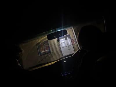 The image, taken at night in Ptichnik, Russia, shows the interior of a car looking out through the windshield. The windshield is wet, possibly from rain, with streaks visible. Directly ahead, through the windshield, is a building with a corrugated metal facade. A white door with a glass window and a silver handle is visible in the center of the building. To the left of the door, a brightly colored poster is affixed to the wall, featuring text and images.

Inside the car, a rearview mirror hangs from the windshield. A digital clock on the dashboard displays "12:45". Below the clock is a car stereo system with illuminated blue buttons. Two figures are partially visible within the car, silhouetted against the outside light. One figure is in the driver's seat on the left, with their head and shoulder obscured by darkness. Another figure is seated in the passenger seat on the right, also mostly in shadow, with their head and upper body visible. The scene suggests the car is parked in front of the building.