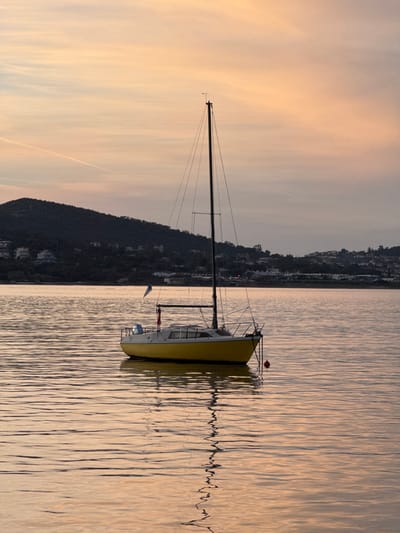 A yellow sailboat rests on calm water reflecting a golden sunset. The mast towers upward, connected by rigging. A small flag flutters on the stern. The water's surface has slight ripples creating a mirrored effect of the boat and sky. A land mass with structures is visible in the background along the horizon, silhouetted against the sky. The sky is a gradient of light oranges and yellows.