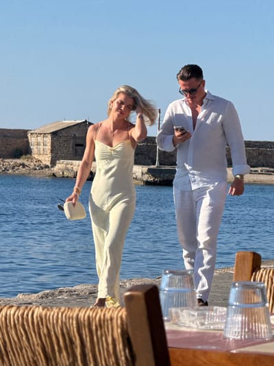 Two people are captured in a vertical photograph, seemingly on a boat or by the water's edge. The man on the left is dressed in a flowing white shirt and pants, with sunglasses perched on his nose. He is looking down at his phone, which he holds in his left hand. A ring is visible on his left ring finger. His posture suggests he is either standing or leaning, with his body angled slightly away from the camera.
To his right, a woman in a pale yellow, spaghetti-strap dress is also visible. Her hair is light blonde and styled loosely. She is holding a small, white clutch in her right hand and appears to be looking down with her eyes closed, one hand resting on her head as if shielding herself from the sun or in a moment of repose. Her left arm, adorned with bracelets, is extended slightly.
The scene is bathed in bright sunlight, indicating a daytime setting, likely during pleasant weather. The background features the calm, blue water of the sea and a stone structure that resembles a building or historical ruins. The water's surface is calm, with a slight ripple effect. In the foreground, part of a wicker-like texture is visible, possibly from outdoor seating or a railing. Two clear glasses, containing a clear liquid, sit on a wooden surface to the right of the man, suggesting they are having drinks. The overall atmosphere is relaxed and luxurious, perhaps a vacation or a special occasion. The location cues, like the stone architecture and the sea, align with the Chania, Greece context. No visible text is present in the image.
