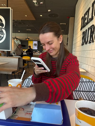 A woman wearing a red and brown striped sweater is seated at a table in a McDonald's restaurant, holding a white smartphone. She appears to be interacting with the phone. On the table are McDonald's food and drink containers, including a cup with a straw. The restaurant interior features black and white patterned seating, a menu board in the background, and other customers. The restaurant is located in the city of Ub, in the Ub Municipality of Serbia.