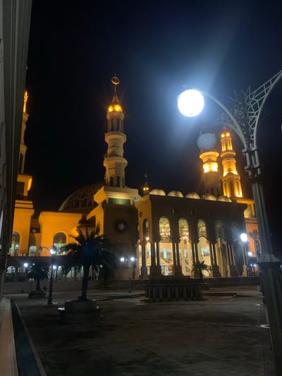 The image presents a grand, illuminated mosque at night, set in Samarinda, Indonesia. The main subject is the mosque's impressive architecture, constructed from light-colored stone or concrete, lavishly lit with warm yellow and white lights that highlight its intricate details.

The mosque features several minarets; one prominent minaret is visible with a golden, crescent moon and star finial. Multiple domes, including a large central dome with elaborate patterns and smaller subsidiary domes, crown the structure. Windows and archways are adorned with decorative screens or patterns, revealing internal illumination and possibly chandeliers.

In the upper left foreground, a geometric, lattice-like structure, likely part of an ornate lamppost or architectural feature, is partially visible. A very bright, circular white light source, appearing to be a powerful floodlight or streetlamp, emanates intense light in this area. Palm trees are seen in the midground and foreground, their dark silhouettes contrasting against the brightly lit mosque and the dark night sky. The ground consists of paved areas, possibly courtyards or pathways.

The scene captures a clear night with no discernible weather events. No people are visible, nor is any specific activity taking place beyond the stationary presentation of the building. The image conveys a sense of peaceful reverence and architectural splendor. No text is visible in the image.