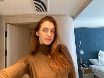 A female individual with reddish-brown hair and light eyes is centered in a close-up, facing the camera directly with a neutral expression. She is wearing a plain, long-sleeved brown turtleneck top. The scene is an interior, indicative of a modern hotel room.

To the subject's left, a light-colored wooden door frame is partially visible, with a slightly ajar door beyond, revealing two dark hinges. Above this, a curved architectural element runs along the ceiling line. The white ceiling at the top of the image features a rectangular air vent with vertical slats.

To the subject's right, a wall clad in vertical blue panels serves as a backdrop. A section of a bed with white linens and pillows is visible on the far right. A light-colored nightstand is positioned between the subject and the bed, holding a black corded telephone, a clear plastic water bottle, a small white cup, and associated charging cables. A wall-mounted lamp with a spherical white fixture extends from the blue paneled wall above the nightstand.

The subject is performing the action of posing for a photograph. No other individuals or direct interactions are visible. The specific location in Licheng District, China, cannot be determined from the visual evidence within the image.