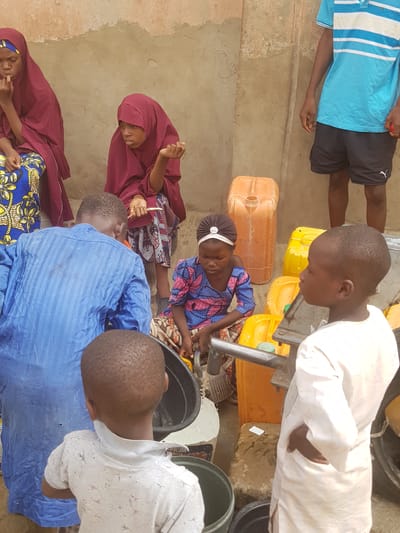 In Dutsinma B, Nigeria, a group of children gathers around what appears to be a water source. Several yellow and orange jerrycans are scattered on the ground. A child wearing a blue and pink patterned dress with a white headband is operating a pump or faucet. Other children watch. Two girls in burgundy hijabs are present. One boy is wearing a blue striped shirt with black shorts. Another child wears a white shirt. A large black bucket is visible.