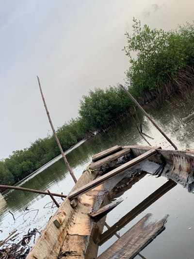 The image depicts a dilapidated wooden structure, possibly a boat or dock, positioned in a body of water surrounded by dense, green vegetation, likely mangroves. The structure is in a state of disrepair with broken planks and debris visible. The water is murky, reflecting the sky and vegetation. The sky appears overcast. Based on the prompt, the location is likely a waterway near Ibasa, Nigeria. There is no visible human presence or activity within the frame.