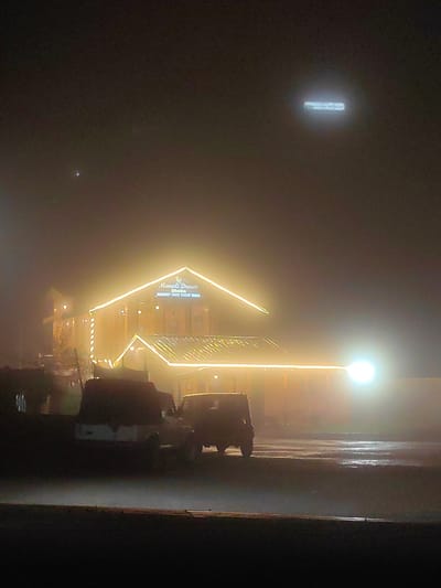 This is a photograph taken at night in Kulang, India, likely during foggy or misty conditions, as evidenced by the diffuse lighting and obscured details. The primary subject is a building, possibly a restaurant or hotel, illuminated by numerous strings of yellow lights that outline its architecture, including the rooflines and vertical elements. A sign above the entrance reads "Manali Dream" and "Dhaba," with smaller text below indicating "BREAKFAST LUNCH SNACKS DINNER." The building appears to be two stories high and has a prominent, well-lit facade.
In the foreground, two vehicles are visible, their forms softened by the low light and atmospheric conditions. One appears to be a pickup truck, possibly with a white canopy, and the other a darker SUV or jeep. Their presence suggests a public space or an establishment accessible by vehicle.
The sky above is dark and hazy, with a bright, slightly diffused light source in the upper right quadrant, which could be a distant light or part of the ambient illumination. There's a small, bright point of light in the upper left quadrant of the sky, possibly a star or planet. The overall atmosphere is subdued and somewhat mysterious due to the darkness and fog, creating a tranquil yet slightly eerie mood. The scene is likely a street or parking area outside the establishment.