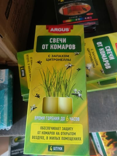 The image displays a close-up view of several boxes of citronella mosquito repellent coils. The primary box in the foreground is predominantly green and yellow, featuring illustrations of grass and cartoon mosquitoes. Text on the box is in Russian, indicating "Coils against mosquitoes" and "with the scent of citronella." Other boxes, partially visible, are stacked behind and to the side of the main box, suggesting a retail or storage setting. The overall scene appears to be indoors, with a wooden surface visible at the bottom and a cardboard box in the upper left corner. The lighting is consistent with indoor ambient light. There are no people or animals present in the image. The specific location in Berdiansk, Ukraine, cannot be determined from the visual information provided.
