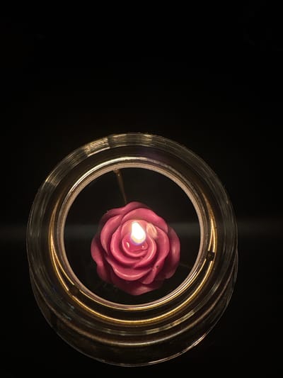 A close-up, top-down shot features a pink rose-shaped candle with a lit wick, floating in water within a clear glass circular container. The container has a decorative, ridged rim with metallic gold accents. The background is entirely black, creating a stark contrast that highlights the candle and container. The scene is devoid of any human presence and is not identifiable as being in Ipswich, United Kingdom.