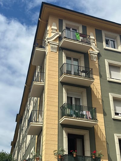 The image shows the exterior of a multi-story building, likely an apartment complex, under a partly cloudy sky. The building has a corner facade with alternating sections of pale yellow and dark green. Decorative architectural elements, such as pilasters and ornate mouldings, adorn the yellow sections. Several balconies with black metal railings are visible along the facade. On the balconies, various items are present, including potted plants, a green towel, a small table and chairs on one balcony, and laundry hanging on a line on another. The windows are predominantly white with closed blinds. In the foreground, the tops of trees can be seen. The style of the building and the presence of balconies are typical of urban architecture in cities like Oviedo, Spain.