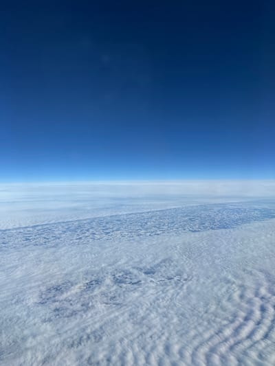 An aerial photograph presents a panoramic view of sky and clouds from a high altitude. The upper half of the image features a clear, deep blue sky, transitioning to a lighter blue near the horizon. Below the horizon, multiple horizontal layers of white to light grey clouds are visible. The middle and lower sections of the frame are dominated by a textured expanse of white and light grey clouds, exhibiting wavy or ripple-like patterns, particularly towards the bottom right. A prominent, dark, elongated band runs horizontally across the mid-cloud layer, revealing patches of the underlying terrain, which appear as mottled dark and light areas, possibly indicating land or snow-covered ground seen through breaks or shadows in the cloud cover. No individuals, identifiable man-made objects, or specific actions are visible within the frame. The location is stated as Dimitrovgrad Municipality, Serbia.