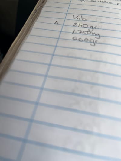 The image shows a close-up of a ruled notebook page with handwritten notes. The notes appear to be in Spanish, with some entries including numerical values followed by units of measurement such as "gr" (grams) and "kg" (kilograms). One entry is labeled "Kilo". There is also a letter "A" written on the left side of the page. The notebook is open, and the binding and edge of a second page are visible on the left. The background is dark and out of focus. There is no discernible information in the image that would indicate a location in Conuco Viejo, Venezuela.