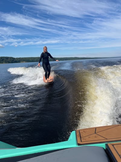 A blonde woman in a black wetsuit is wakeboarding on a dark body of water under a blue sky with wispy white clouds. She is riding on a light orange wakeboard, balanced with her arms extended to the sides for stability. The wakeboarder is positioned on a wave created by a boat, which is partially visible in the foreground, showing its turquoise railing and a brown deck with a geometric pattern. The background features a dense green forest line bordering the water, indicative of a natural landscape. The image captures a moment of athletic activity on a body of water, likely a lake or river, under daylight conditions. The location is identified as Gromovo, Russia.