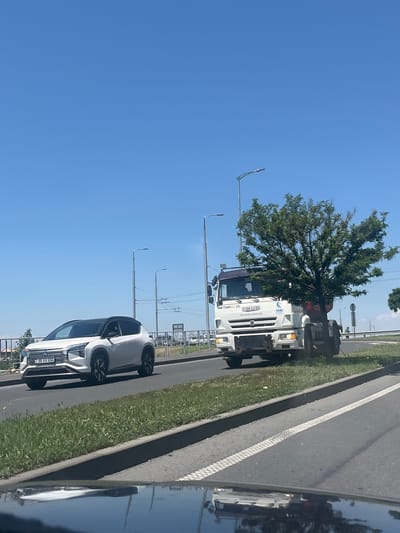 A white Mitsubishi Outlander SUV with license plate "55 VV 050" drives on a highway in Mkhchyan, Armenia. To its right, a white Kamaz truck with license plate "684 LU 01" is parked on the side of the road, partially obscured by a large tree. The sky is clear and blue, and the road is bordered by green grass. The scene appears to be captured from another vehicle moving on the highway.