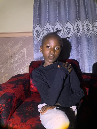 The image shows a young Black child lying on a patterned sofa in what appears to be an indoor setting. The child is wearing a black long-sleeved shirt and has their arms crossed over their chest. Their gaze is directed towards the camera. A white object, possibly a pillow or blanket, is visible next to them, partially obscuring their body.
The sofa has a dark red base with a black floral pattern. Behind the child, a curtain with a grey and white floral design is visible. The lighting suggests it might be evening or the room is dimly lit. The background wall is a light beige color with some white splatters or marks.
The image is oriented vertically, with the child's head appearing at the top left of the frame. The child's expression is neutral. No text is visible in the image. The location context is Barakin Sabawa, Nigeria.
