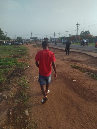 The image shows a man walking along a dirt road. He is wearing a red shirt and blue shorts, and walking in white and black sandals. He is walking away from the camera, and his head is down. There are power lines running overhead, and a few cars and motorcycles can be seen in the distance. There is green vegetation on either side of the road. The scene is in the city of Kanshio, Nigeria, located at the coordinates 7.717° N, 8.543° E.