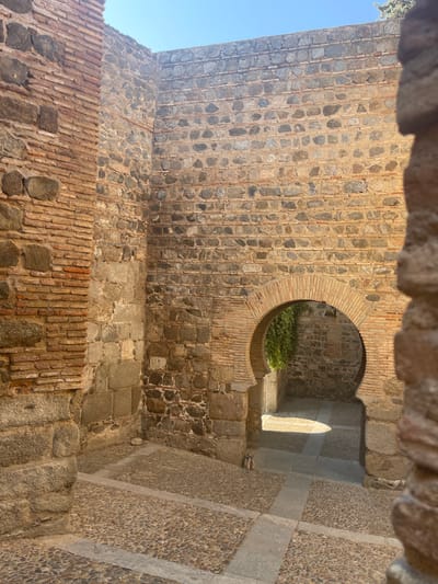 The image shows a view of an arched doorway in Toledo, Spain, specifically within the historic Jewish Quarter. The doorway is constructed from brick and leads into an area with lush green vegetation. The walls surrounding the doorway are made of stone and brick, showcasing the architectural style of the region. The ground is paved with cobblestones and stone slabs, with sunlight casting shadows on the path. The sky is clear and blue, indicating fair weather. There are no people visible in the image.
