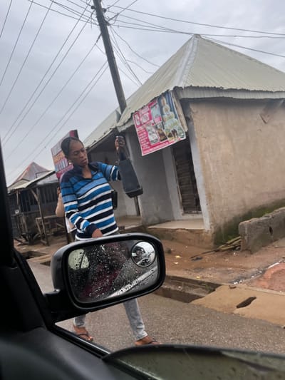 A woman wearing a blue and white striped long-sleeved shirt and gray pants walks on a wet road, holding a black bag in her right hand. She is seen in the foreground, with a car's side mirror on the left side of the frame, showing a reflection of the woman and a motorcycle. In the background, there is a building with a corrugated metal roof, a utility pole with numerous wires, and other structures that appear to be shops or small dwellings. The scene is suggestive of a street in a developing urban area, possibly Sabojibi, Nigeria, given the presence of the utility pole, the style of the buildings, and the overall atmosphere. The ground is wet, indicating recent rainfall.