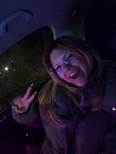 The scene is located in the city of Skalsko, Bulgaria. The image captures a young female individual seated inside a vehicle at night. She is positioned towards the left of the frame, smiling broadly with her mouth open, revealing her teeth. Her hair is light brown, framing her face and resting on her shoulders. She wears rimless eyeglasses, the lenses reflecting multiple distinct blue and purple light sources. She is dressed in a dark, possibly padded, jacket. Her right hand is raised, forming a "V" or peace sign with her index and middle fingers; her fingernails are long and appear to have white tips. The vehicle's interior is dimly lit with a predominant purple and blue hue. Visible elements include a light-colored grab handle on the dark roof lining in the upper left corner and dark seat upholstery. Through the vehicle's window on the left, the dark exterior environment shows faint, out-of-focus yellow-orange light sources.
