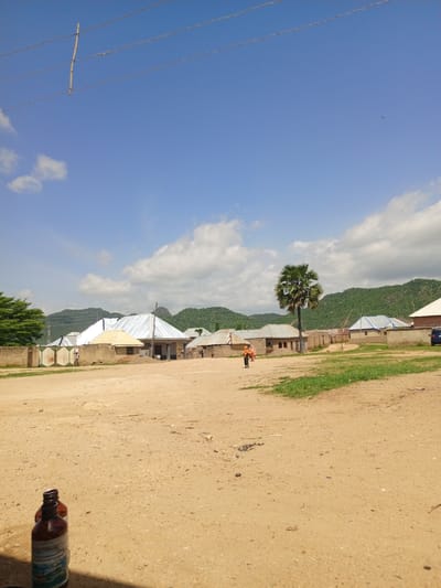 The media file captures a scene in Barakin Babele, Nigeria, under a bright, partly cloudy sky, suggesting it is daytime. The setting is a village or rural area with several buildings visible. These structures appear to be houses with corrugated metal roofs, some with light-colored walls and others with darker, possibly brick or mud, construction. A tall palm tree stands prominently in the middle ground.
In the foreground, a dusty, unpaved ground dominates the lower portion of the image, with a few small stones and debris scattered across it. Two brown glass bottles, one partially obscured by shadow, are positioned in the bottom left corner.
Further into the scene, a few people are visible. One person, dressed in light-colored clothing, stands near a gate in the left mid-ground. Further to the right, two individuals, one appearing to be a woman in a headscarf and the other a child, are walking across the open area.
The background features rolling green hills or mountains, with a line of trees along their slopes. Power lines are strung across the sky, supported by a wooden pole in the upper left quadrant. The overall impression is of a typical day in a rural Nigerian community, with daily life unfolding against a backdrop of natural scenery and modest architecture. No specific activity or event is clearly depicted beyond the presence of people moving about.