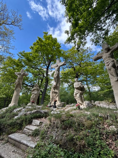 The image depicts a religious scene set in the town of Častá, Slovakia. Three stone sculptures depicting the crucifixion of Jesus Christ are positioned on a rocky, overgrown hillside. The sculptures are arranged vertically, with the uppermost sculpture partially visible at the top edge of the image, the middle one fully visible, and the lowermost sculpture partially visible at the bottom. Each crucifixion scene includes a cross and at least one additional figure, presumably mourners. Green trees and a blue sky with some clouds form the backdrop. The perspective is from below, looking upwards towards the sculptures. Some flowers have been placed next to the sculptures. A vapor trail from an airplane is also seen.