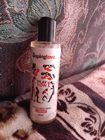 A bottle of "Dopinglove LUBRICANT" is pictured, placed on a patterned blanket. The bottle is white with black text and red accents, featuring an illustration of figures. The text indicates "PLAY NOW" and "for all types of love," with a volume of 200 ml. The background consists of a textured, multi-colored fabric. The image appears to be taken indoors. The bottle is upright and in focus.