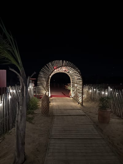 The image depicts an illuminated archway made of natural wood slats on a sandy beach at night, located in Torreilles, France. A wooden boardwalk leads through the arch, which is adorned with string lights. To the left, a palm tree stands beside a rustic wooden fence also decorated with string lights. A woven lantern sits on the sand near the boardwalk, and a potted plant is visible to the right. The background is dark, suggesting nighttime, with a dimly lit structure visible through the arch. The overall scene conveys a festive and inviting atmosphere.