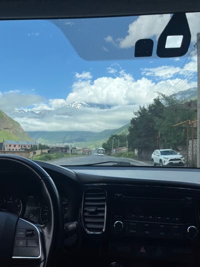 The image is taken from inside a car, looking out through the windshield. The view shows a road in a mountainous area. The road curves to the right, with trees and buildings lining its sides. In the distance, snow-capped mountains rise against a blue sky with scattered white clouds. A white SUV is parked on the right side of the road. The interior of the car is visible, including the steering wheel, dashboard with a digital display showing "TIRE PRESSURE MONITOR SERVICE REQUIRED", and the car's entertainment system. The location is identified as Arsha, Georgia. The scene depicts a journey through a scenic landscape.
