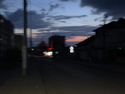 The image depicts a street scene in Topolovgrad, Bulgaria at dusk. The sky shows a mix of dark clouds and a hint of sunset colors. On the left, a residential building is partially visible. A utility pole stands near the sidewalk with posters on it. Across the street, a brightly lit shop or establishment contrasts with the surrounding darkness. Further down the street is a building with what appears to be a red neon sign. A road sign with a white diamond with an arrow indicating a turn to the right. On the right side of the street, a dark building is situated. The street itself is dark and appears wet.