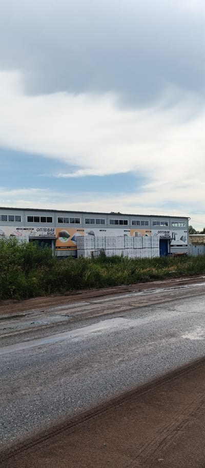In Pirogovo, Russia, a long, grey industrial building with numerous windows lines the horizon under a cloudy sky. The building features a prominent banner that reads "ОПТОВАЯ БАЗА" (Wholesale Base). In front of the building, stacks of white, shrink-wrapped materials, possibly construction supplies, are visible. Adjacent to these stacks, a dense area of green vegetation and grass grows. The foreground of the image shows a dirt and gravel road with puddles of water, suggesting recent rain. Tire tracks are visible on the road surface. The overall scene depicts a commercial or industrial area on an overcast day.