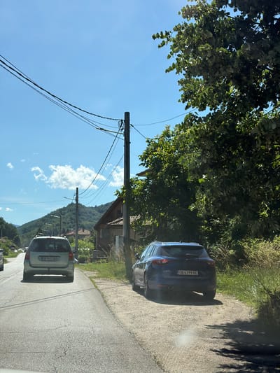 The image depicts a sunlit day in Beli Osam, Bulgaria, featuring a rural road scene. A light-colored station wagon with license plate OB 9023 BH is traveling away from the camera on the left side of the asphalt road. To the right of the station wagon and slightly ahead is a dark blue SUV, also traveling away from the camera, with license plate OB 6399 BE. Both vehicles appear to be in motion.
The road is bordered by a dirt shoulder on the right, with abundant green vegetation and tall grass. Several utility poles with numerous wires are visible along the roadside, crossing overhead. In the background, residential buildings with visible roofs are nestled amongst trees and a hillside covered in dense green foliage. A prominent, large tree with lush green leaves dominates the right side of the frame, casting a shadow on the road. The sky is a clear blue with a few scattered white clouds. The overall impression is a bright, summery rural landscape.