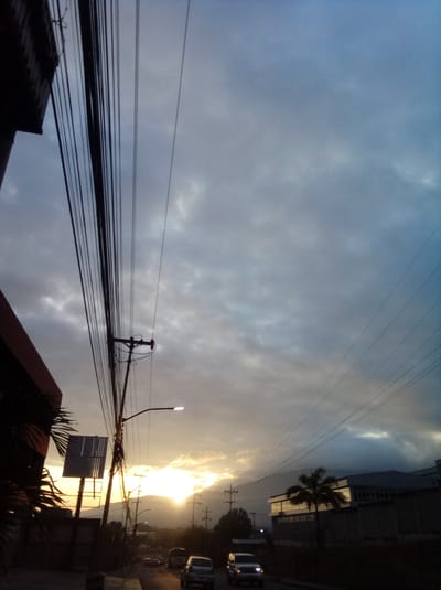 The image shows a street scene in Parroquia Guatire, Venezuela, at what appears to be sunset or sunrise. The sky is filled with clouds, with the sun positioned low on the horizon, partially obscured by hills or mountains in the distance. Power lines and utility poles are prominent in the foreground, running across the sky. A street light is visible on the left side of the image. On either side of the street are buildings and some foliage. Cars are driving on the right side of the image. The overall lighting is dim and shadowy.