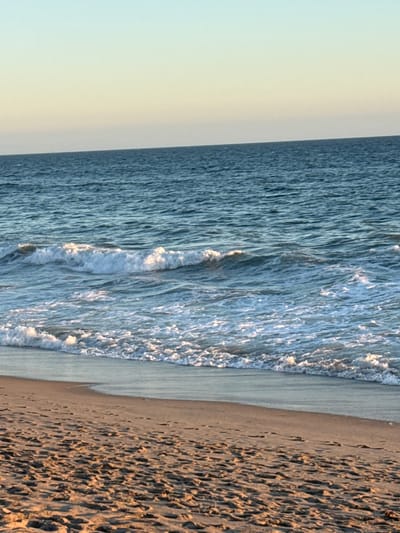 The image depicts a tranquil beach scene at dusk in Malibu, California. The foreground is dominated by soft, sun-kissed sand, imprinted with numerous footprints, suggesting recent human activity. The sand transitions to the gentle, lapping waves of the Pacific Ocean. The water exhibits a deep blue hue, with white foam cresting on the incoming waves, creating a dynamic yet soothing visual. The horizon is a sharp line where the ocean meets a pale, gradient sky, transitioning from a soft yellow near the horizon to a clear, light blue at the top of the frame. There are no people or specific landmarks visible, emphasizing the natural beauty of the coastline.
