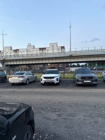 The image shows a parking lot filled with several cars. In the foreground, a black car's front right fender and hood are visible. Behind it, a silver Hyundai Sonata is parked, displaying a license plate that reads "B672TK 790". To its right is a white Range Rover Evoque, with a license plate "E856BY 250". Further to the right is a dark gray Geely Monjaro, its license plate visible as "E991KK 797". In the background, there is a large elevated structure, likely a bridge or highway overpass, with modern buildings behind it. The sky is partly cloudy with a pale blue hue. The ground is covered in gravel. The scene is set in a developed urban or suburban area, consistent with the city of Voyennosluzhashchiy, Russia. There are no people or animals visible in the image.