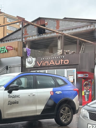 This is a vertically oriented, outdoor photograph taken in Фролы, Russia, likely during the day under overcast skies, as suggested by the diffused light and the wet pavement. The primary focus is on a white car with blue and grey accents, part of a ride-sharing service, parked in front of a commercial building. The car's side displays text in Russian, "Яндекс Драйв" (Yandex Drive), indicating its affiliation.
The building behind the car appears to be under renovation or in a state of disrepair. It has a dark brown, corrugated metal facade with a prominent sign that reads "VinAuto АВТОЗАПЧАСТИ" (VinAuto Auto Parts) in large, orange, illuminated letters. Below this, a circular logo with a vehicle silhouette is visible, also with "VinAuto" inscribed. A red vending machine, labeled "ROCKY BOXER" and with "RB" logos, is situated to the left of the "VinAuto" sign. Further to the left, a sign with green and red lettering, partially visible, reads "КУНГУРСКИЙ" (Kungurskiy), which likely refers to a local product or business.
The scene suggests a commercial area, possibly a street or parking lot, with elements of commerce and transportation. The wet appearance of the ground implies recent rain. No people are clearly visible, though shadows suggest some presence within the building or on the street. The overall impression is one of everyday urban activity, with a focus on vehicles and local businesses.