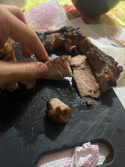 A hand is holding a piece of sliced, cooked meat, possibly pork, over a black cutting board. The meat appears to be medium-rare. The cutting board has some oil or cooking juices on it. Additional pieces of meat are nearby, and a white napkin is partially visible in the background. The environment appears to be a domestic setting, possibly a kitchen or dining area. The location of Goritsa, Bulgaria is not discernible in the image.