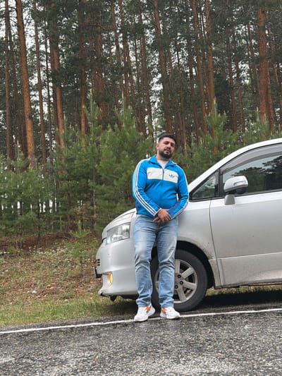 A man wearing a blue and white track jacket and jeans is leaning against the driver's side of a silver van parked on the shoulder of a road in a wooded area. The road is asphalt with a white line marking the edge. The trees in the background appear to be pine trees, indicating a forest-like environment. Based on the provided location of Сизая, Russia, the image likely represents a roadside scene in this region. The interaction is between the man and the vehicle in a natural environment near a road.
