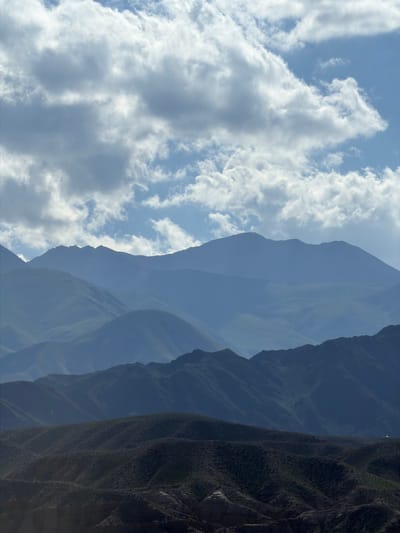 The image is a vertical, low-angle shot of a vast, mountainous landscape. The left side of the frame features a bright blue sky filled with fluffy white clouds, suggesting daylight. The clouds are illuminated by sunlight, casting subtle shadows and adding texture to the sky.
Moving towards the right, the scene transitions into layers of mountains. The closest mountain range appears as a dark, shadowy form, with visible texture suggesting steep slopes and perhaps vegetation or rocky surfaces. Beyond this, successive mountain ranges fade into softer shades of blue and purple due to atmospheric perspective, creating a sense of immense depth and scale. The distant peaks are less defined, blending with the sky and creating a hazy, ethereal quality.
The overall impression is one of a majestic and remote natural environment. The dramatic layering of mountains and the contrast between the bright sky and the shadowed peaks evoke a feeling of awe and grandeur. The image captures a moment of natural beauty, with no discernible human presence or activity. The time of day is likely daytime, possibly late morning or early afternoon, given the bright sunlight. The weather appears clear with scattered clouds.