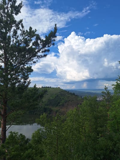 The image depicts a landscape view from a hill overlooking the city of Sim, Russia. In the foreground, a large pine tree is prominent on the left, with its branches extending into the frame. The middle ground features a body of water, likely a river or lake, reflecting the sky. Beyond the water, a green, rolling hill rises, dotted with evergreen trees and a path winding its way up. In the distance, the city of Sim is visible, with buildings and structures nestled within a valley. The sky is dramatic, with a mix of bright blue patches and large, white, cumulus clouds. Towards the horizon, darker clouds suggest an impending rain shower. The overall impression is one of a natural landscape meeting a populated area under a dynamic sky.