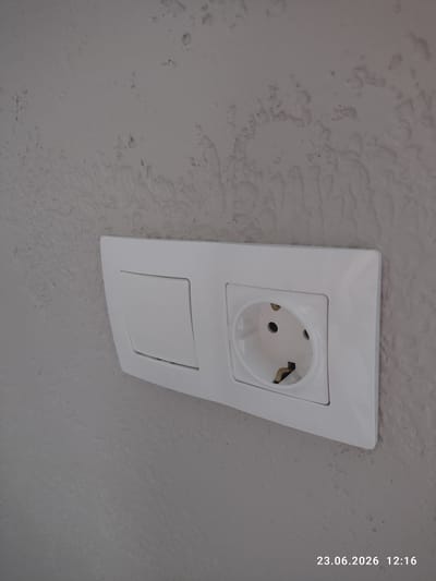 The image shows a white electrical outlet and a blank plate installed on a textured, light gray wall. The outlet is a European-style socket with three round holes. The blank plate is rectangular and positioned below the outlet. The wall has an uneven, bumpy texture. The timestamp "23.06.2026 12:16" is visible in the upper right corner of the image, suggesting it was taken in the early afternoon. There are no people or specific weather conditions depicted. The setting appears to be indoors, likely a residential or commercial building. The overall scene is mundane, focusing on architectural elements.