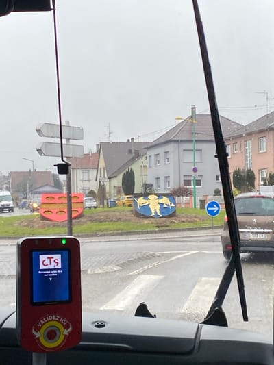 The image shows a view from inside a bus or tram. The ticket validation machine is visible, displaying the logo of "CTS" and text in French. Through the windshield, a street scene is visible on a rainy day. There are signs, buildings, and other street furniture. There are also vehicles. The scene is typical of a European town or city.