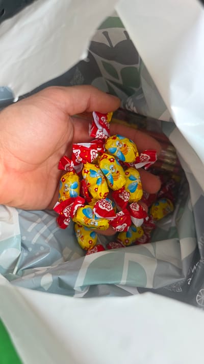 A close-up shot from above shows a person's hand holding a pile of individually wrapped candies. The candies are oval-shaped and feature a yellow wrapper with a cartoon bee and red flowers. Some of the wrappers are red with white writing. The candies are inside a white bag with a gray and light blue patterned lining. The bag appears to be a reusable shopping bag. The image is taken from a high angle, looking down into the bag. The location is not specified in the image or any provided context.