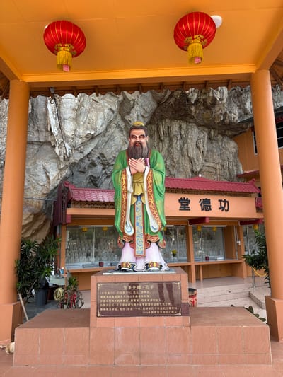 In Ipoh, Malaysia, a statue of Confucius stands prominently in front of a building with a prominent sign displaying Chinese characters. The statue depicts Confucius in traditional green and gold robes, with a long beard and clasped hands. The building behind the statue has an orange facade and a red tiled roof, with several windows displaying what appear to be historical artifacts or religious items. The scene is set against a backdrop of a large, rocky cliff face. Two large red lanterns hang from the ceiling above the statue, adding a festive touch. The ground in front of the statue is paved with terracotta tiles. A small dog is visible lying on the tiled ground to the left of the statue. The overall atmosphere suggests a place of historical or cultural significance, possibly a temple or museum.