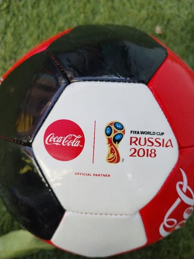 A soccer ball rests on a green grassy surface in Sucina, Spain. The ball features a multi-paneled design in red, black, and white. A prominent white hexagonal panel displays the red Coca-Cola logo within a red circle, a vertical red line, and the FIFA World Cup Russia 2018 logo with accompanying text. Below these, the words "OFFICIAL PARTNER" are printed. Additional white stylized swirl patterns are visible on a red panel to the right. The ball exhibits visible stitching along its panel seams. No individuals are present, and no actions or interactions are depicted.