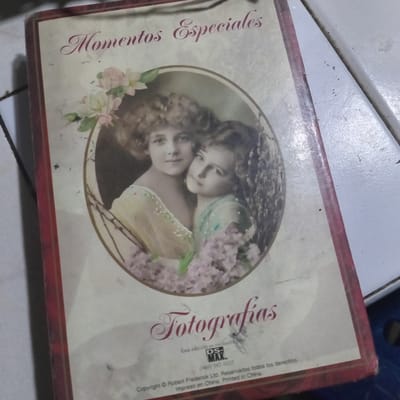 The image shows the cover of a photo album. The title "Momentos Especiales" (Special Moments) is written in cursive at the top. Below the title is a framed oval photo featuring two young girls with curly hair. The girl on the left is wearing a light-colored dress with floral accents, and the girl on the right is wearing a light blue dress. They are surrounded by flowers, with pink and white blossoms framing the top left and purple blossoms at the bottom. The word "Fotografias" (Photographs) is written in a large, decorative script at the bottom of the cover. Below this are the words "Esta edición es exclusiva para DS-MAX" (This edition is exclusive to DS-MAX), followed by a phone number "(949) 587-9207". The bottom of the cover also contains copyright information: "Copyright © Robert Frederick Ltd. Reservados todos los derechos. Impreso en China. Printed in China."

The photo album is lying on a white surface, possibly a table or countertop, with some textured areas and a dark shadow on the left. There are specks of dirt and some scuff marks visible on the surface and the album cover. The overall impression is that the photo album is an older item, possibly a keepsake or a product from a specific company. There is no specific indication of the location being in Duaca, Venezuela, or any other specific geographical location.