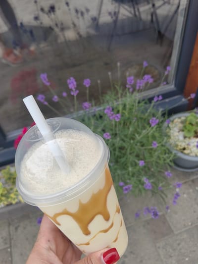 A close-up shot captures a hand holding a clear plastic cup filled with a creamy, light-colored beverage, possibly a milkshake or smoothie, drizzled with a darker caramel sauce. A white straw is inserted into the cup. The background is slightly blurred, showing a gray paved area, a window reflecting outdoor elements, and several sprigs of lavender with small purple flowers. A potted plant with light-colored gravel and green foliage is visible to the right. The scene appears to be outdoors, possibly near a cafe or shop entrance in Slavyansk-na-Kubani, Russia.