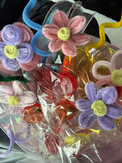 The media file shows a close-up view of a bouquet of pipe cleaner flowers and some translucent, colorful candy or gummy decorations. The flowers are various pastel colors, including pink, purple, and light yellow, and are wrapped in clear plastic. Some of the gummy decorations are red and orange, appearing to be heart-shaped or circular. The bouquet appears to be a gift or decoration. There are no people visible, and no specific time of day, weather, or location cues are apparent beyond the items themselves. There is no visible text in the image.