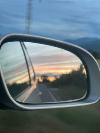 The image captures a view through a car's side mirror, reflecting a sunset over a road. The sky displays a gradient of blue, orange, and pink hues from the setting sun. Trees and hills are visible in the background. A portion of the car's body is reflected in the mirror on the left side. The road appears to be asphalt and stretches into the distance. A blurred telephone pole is visible in the upper left corner of the main image. The setting is rural, consistent with the location of Dabnitsa, Bulgaria, a village characterized by its natural landscapes and agricultural surroundings.