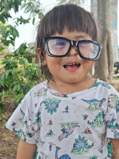 A young girl wearing large black-framed glasses and a white t-shirt with a colorful dinosaur print is pictured from the chest up. Her mouth is open, revealing her teeth and tongue. The girl has dark hair that is slightly messy. The image is taken outdoors, with blurred green foliage in the background and a rustic, wooden-like structure above. The lighting suggests it is daytime, and the weather appears to be clear. The overall mood is playful and spontaneous. No visible text is present. The setting is likely a garden or a natural outdoor space.