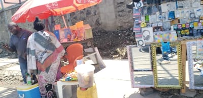 The scene is a street vendor's stall in Rubavu, Rwanda. The stall is located at coordinates latitude: -1.668, longitude: 29.268. The vendor is selling a variety of items, including headphones, mirrors, and other small goods. There are two people at the stall, one is a man standing to the left of the stall, looking at his phone, and the other is a woman sitting at a table in front of the stall. The man is wearing a dark grey shirt. The woman is wearing a white and red patterned shirt. The stall is set up against a wall and there is a large red umbrella over it. The ground is paved. There is a bright yellow bucket to the right of the stall. The sun is shining and there are shadows cast from the umbrella and the wall. There is a plastic container full of a brown substance, likely a snack,  on the table next to the woman. There are orange and white chips stacked to the left of the container. There is also a small blue cooler to the left of the woman. There are four mirrors, two of which are framed in gold, that are propped up on the stall. The mirrors reflect the scene behind them.