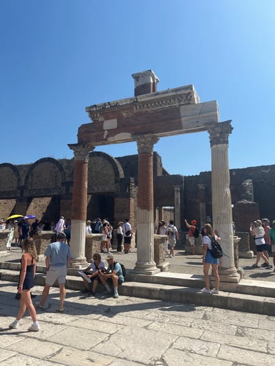 The image depicts a sunny day in the ancient city of Pompeii, Italy, likely within the Forum. The foreground is dominated by a large, partially ruined stone structure with imposing brick columns supporting a weathered archway. The pavement is made of large, irregular flagstones.

In the immediate foreground, a woman with curly blonde hair, wearing a dark blue slip dress and white sneakers, is walking towards the left. Behind her, a man in a light blue t-shirt and grey shorts stands with his back to the camera. To his right, two people, a woman and a man, are seated on stone steps, the woman holding a map and the man looking at his phone.

In the mid-ground, a group of tourists are visible. Some are standing, observing the ruins, while others are walking. Several people are holding umbrellas, suggesting a hot day. The background shows the well-preserved walls of ancient Roman buildings, characterized by brickwork and arched openings, typical of Pompeii's archaeological site. The sky is a clear, bright blue.